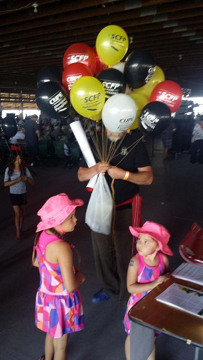 Man holding red, yellow, red and black balloons with two little girls in pink dresses and hats.