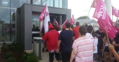 The backs of a crowd of men and women holding placards and flags, on the sidewalk in front of and entering the doorway of a grey building.