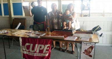 Two men and a woman holding a baby behind a wooden table with a pink flag saying CUPE on the front