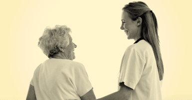 Young women with long hair wearing hospital scrubs holds the arm of an elderly woman, both smiling