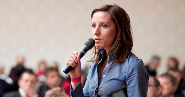 30ish white woman in a blue shirt holding a microphone and speaking