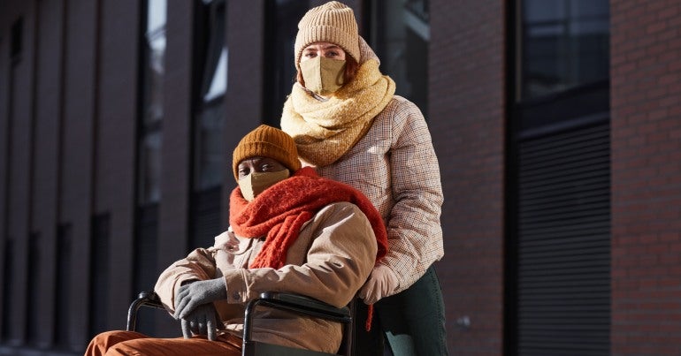 Man in wheel chair with woman standing behind him both wearing masks and winter clothes