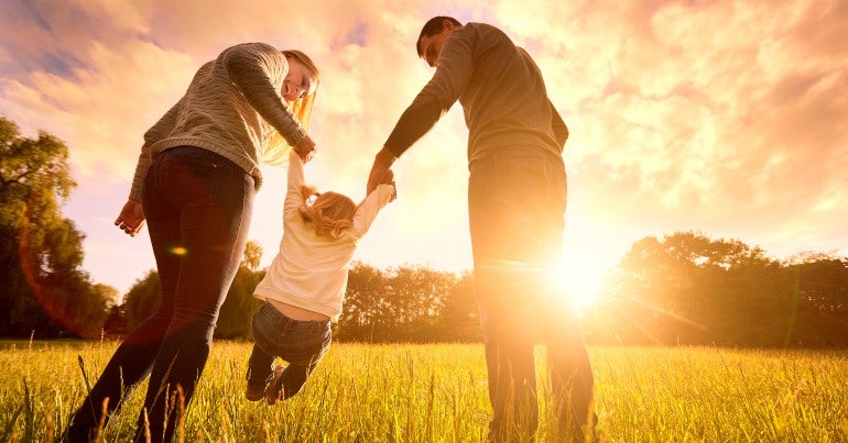 A child holds two adults' hands is swung between them. In the background the sun is low in the sky.