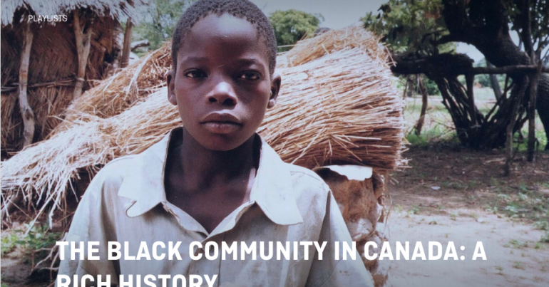 A young Black boy looks directly out of the frame, there are trees and a bundle of straw behind him