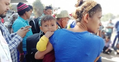 Picture shows a crowd of people, a woman in the foreground looking to the side, a child on her hip looks straight out at the viewer