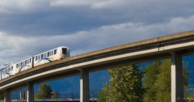 Image of Vancouver SkyTrain, elevated light rapid transit train and track with mountains and blue sky in the background.