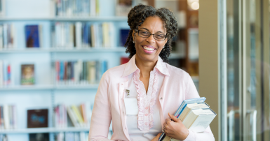 A cheerful mature female librarian stands in her library and smiles for the camera.  She holds a stack of books as she laughs.