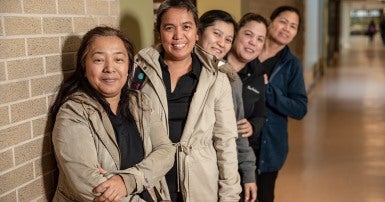 Group of five smiling women in line against a wall, leaning out to show their faces