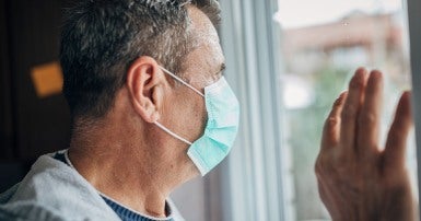 Elderly man wearing blue face mask looking out a window, hand resting on window