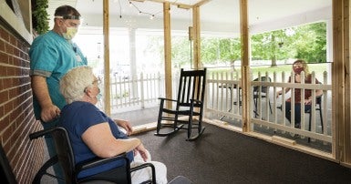 Long-term care worker in a mask and face shield stands next to an elder mask-wearing woman sitting in a wheelchair, speaking to a masked woman sitting outside a screened-in porch.