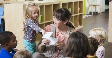 Child care worker surrounded by children, handing something to a girl with Down syndrome