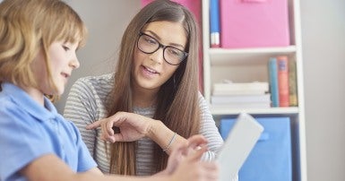 A short haired blond child is looking at an electronic tablet, a long haired woman with glasses also looks at the tablet