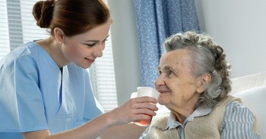 Young woman in blue scrubs standing beside bed holding a cup of orange liquid to the mouth of an elderly woman 