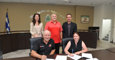 Two people sitting at a table with signed contracts and three people standing behind them 