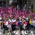 Very large crowd with pink flags in a city street, police and bicycles in the foreground.
