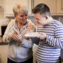Middle aged woman holding a phone, young man eating a piece of pizza, both standing in a kitchen