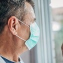 Elderly man wearing blue face mask looking out a window, hand resting on window