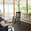 Long-term care worker in a mask and face shield stands next to an elder mask-wearing woman sitting in a wheelchair, speaking to a masked woman sitting outside a screened-in porch.