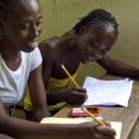 Two Black women at a wooden desk writing with pencils on pads of paper