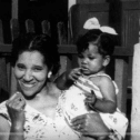 Black and white photo of a smiling black woman holding a black toddler girl in her arms, standing in front of a porch