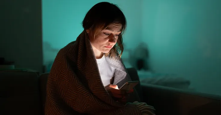 Person wrapped in blanket in a dark room looking at their phone
