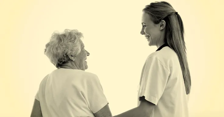 Young women with long hair wearing hospital scrubs holds the arm of an elderly woman, both smiling