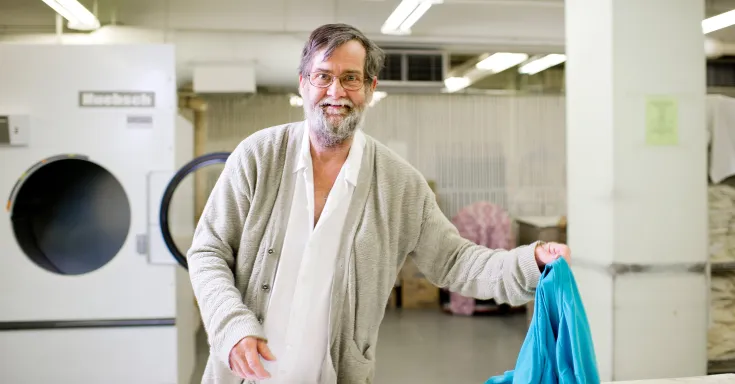 Bearded man holds a turquoise garment. Behind him is an industrial washer.