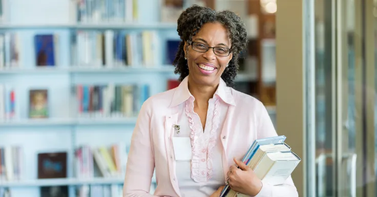 A cheerful mature female librarian stands in her library and smiles for the camera.  She holds a stack of books as she laughs.