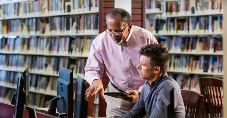 Two multi-ethnic men in the library looking at a computer screen and conversing. A mature man in his 40s is sitting at the desktop PC and a senior African-American man in his 60s is standing beside him.