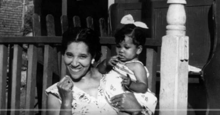 Black and white photo of a smiling black woman holding a black toddler girl in her arms, standing in front of a porch